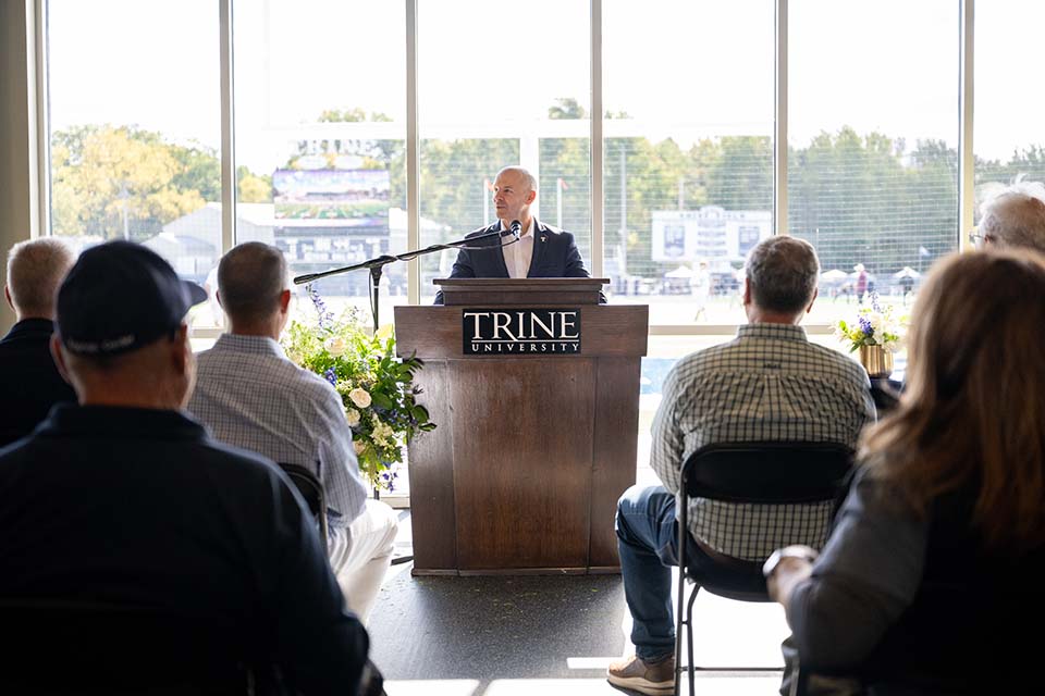 Dr. Kline at podium speaking to crowd. Behind him, outside windows, is the football field